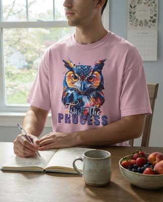Someone wearing a light pink owl t-shirt sits at a kitchen table journaling beside a ceramic mug.