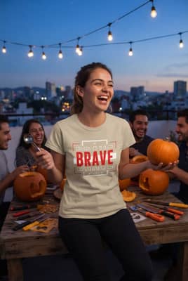 Woman laughing at a rooftop pumpkin-carving hangout at dusk, string lights overhead and the “Be Brave” tee clearly visible with a matte print.