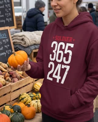 A young woman picks out a small pumpkin at an outdoor farmers market stall on a cloudy morning, wearing a maroon motivational hoodie.