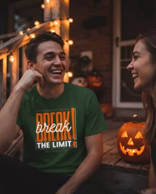 Person in a printed tee laughs with a friend on a dim porch beside string lights and a glowing jack-o'-lantern.