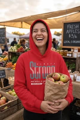 Person wearing a red hooded sweatshirt standing at a farmers market holding a paper bag of apples.