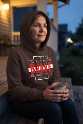 A woman in her early sixties sits on a porch step at dusk, holding a mug with her hood raised against the wind.