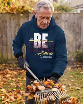 A man in his late sixties rakes leaves in a bright backyard with his hoodie sleeves pushed up to show ribbed cuffs.