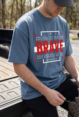 A person wearing a t-shirt sits on the open tailgate of a pickup truck at a forest trailhead while tying their hiking boots.