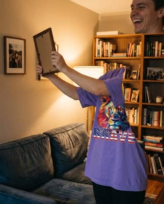 A person in a violet t-shirt smiles while adjusting a framed picture on the wall of a warmly lit living room at night.