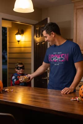 Person wearing a navy t-shirt standing at a kitchen counter handing candy to a trick-or-treater at an open front door.