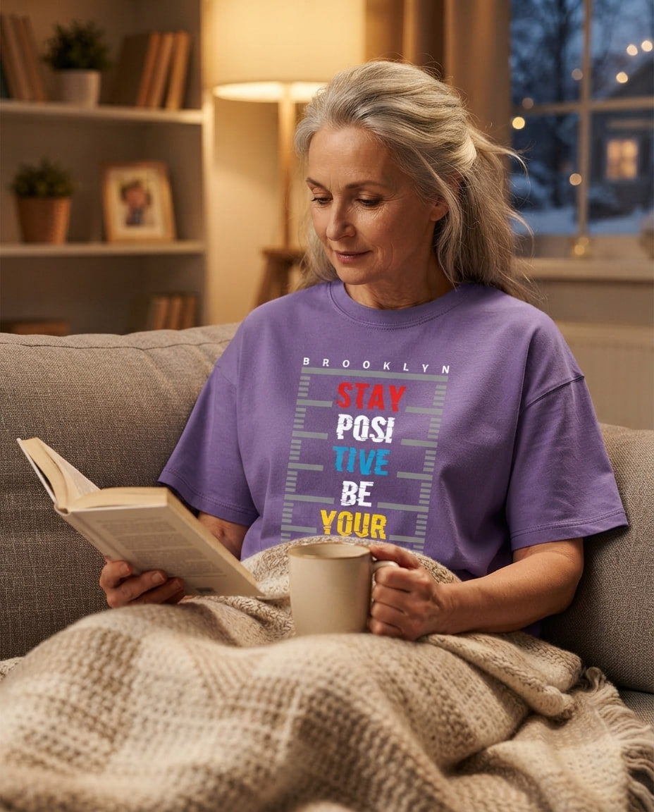 Woman in her fifties reads on a sofa with a mug and blanket, wearing a visible graphic tee under lamplight.