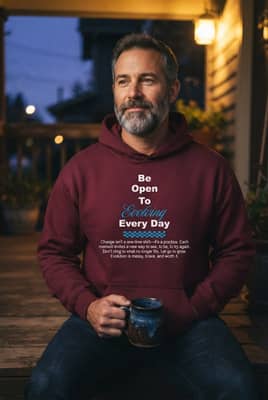 A man wearing a maroon printed hoodie sits on a front porch step at dusk with a mug and his hands in the pocket.