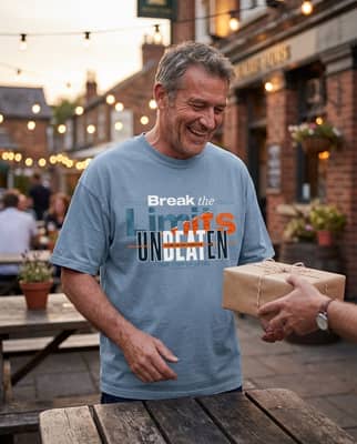 Man in his fifties laughing as a friend hands him a wrapped gift at a pub outdoor table under string lights at dusk.